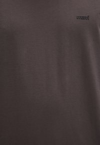 Dunkelbraunes T-Shirt aus weichem Baumwollstoff, mit einem kleinen, schwarzen, geprägten "mavi"-Logo auf der Vorderseite. Schlichtes, unifarbiges Design.