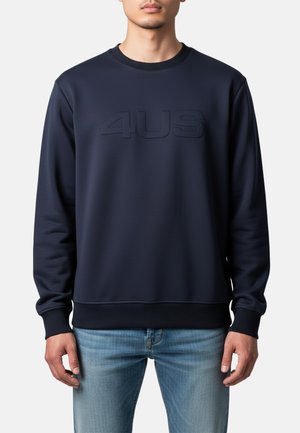 Uomo che indossa una felpa blu navy con la scritta "4US" in rilievo sul petto e jeans azzurri, in piedi davanti a uno sfondo semplice.
