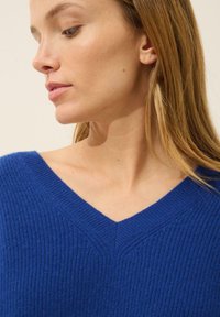 Blå v-ringad stickad tröja med ribbad textur. Har en bred halsringning och subtila sömdetaljer längs axlarna. Mjuk material.