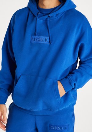 Osoba ubrana w niebieską bluzę z kapturem SikSilk i dopasowane spodnie, z rękami w przednich kieszeniach na białym tle.