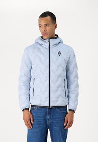 Giacca puffer blu chiaro con un motivo trapuntato a zigzag, zip nera, cappuccio e un piccolo logo sul petto. Abbinata a jeans blu.