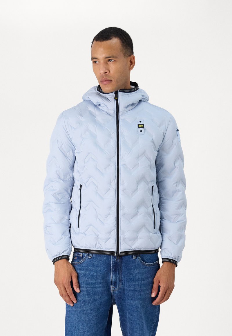 Giacca puffer blu chiaro con un motivo trapuntato a zigzag, zip nera, cappuccio e un piccolo logo sul petto. Abbinata a jeans blu.