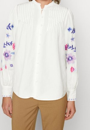 Witte blouse met lange mouwen, knopen aan de voorkant, geplooide schouderpas en kleurrijke paarse en roze bloemenborduursels op de mouwen.