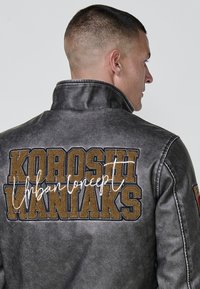 Chaqueta de cuero gris con un alto cuello, bordado "KOROSHI WANIAX" en marrón y "Urban concept" en blanco en la parte posterior, acabado texturizado.