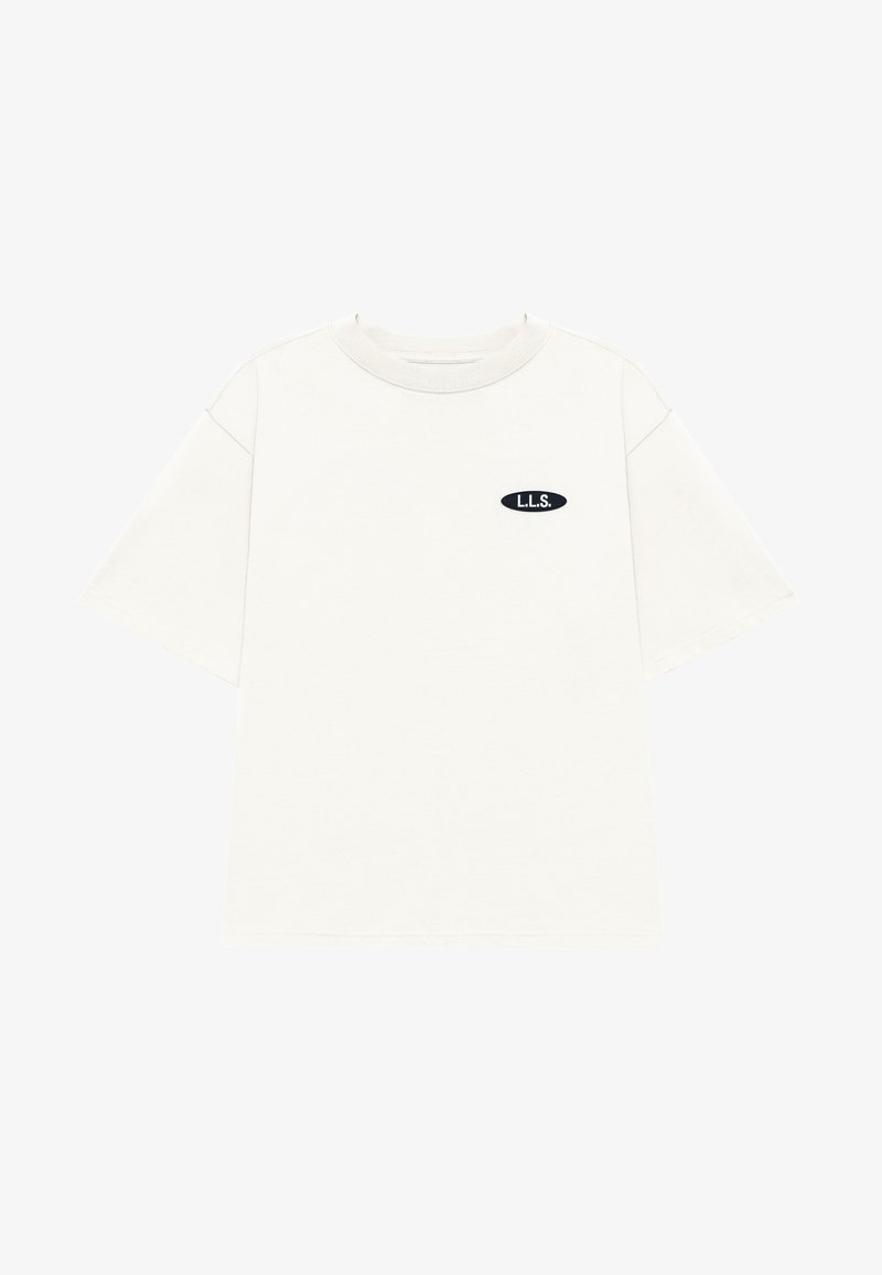 T-shirt à manches courtes et col rond en tissu beige clair. Présente un petit patch ovale noir avec le logo "L.L.S." sur la poitrine.