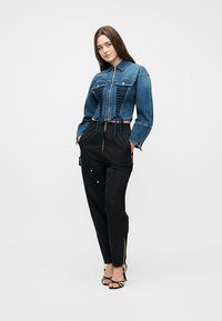 Γυναίκα όρθια με τα χέρια στις τσέπες μαύρου παντελονιού, φορώντας μια εφαρμοστή μπλε τζιν ζακέτα με δέσιμο μπροστά και μαύρα πέδιλα με λουράκια σε λευκό φόντο.