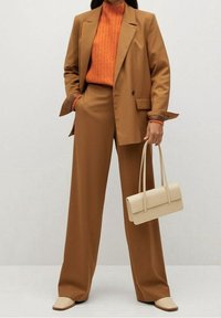 Costume marron sur mesure avec pantalon large, pull à côtes orange et sac à main beige. Tissu lisse, design structuré et accents minimalistes.