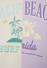 T-shirt de algodão em creme e lavanda, com design tropical que inclui palmeiras, pássaros e acentos florais em turquesa e amarelo.