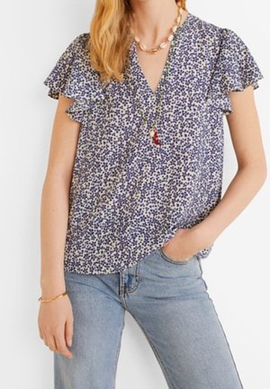 Femme portant un chemisier à manches courtes à fleurs bleues et blanches avec des manches à volants et un jean bleu clair, accessoirisée d'un bracelet doré et de colliers superposés.