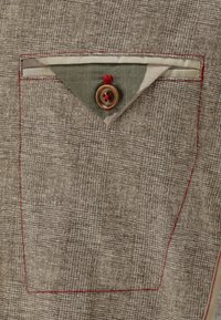 Il tessuto marrone con trama presenta una tasca a toppa triangolare con motivo mimetico, rifinita da cuciture rosse e un bottone marrone.