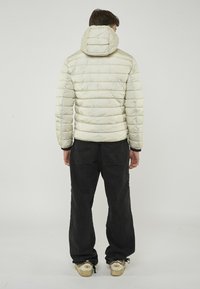 Uomo di spalle che indossa un piumino leggero color beige con cappuccio, pantaloni neri e sneakers beige, su uno sfondo chiaro uniforme.