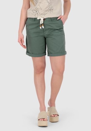 Frau trägt grüne Shorts mit aufgerolltem Saum und Kordelgürtel, beiges T-Shirt mit Grafik und beige Plateausandalen, steht mit einer Hand in der Tasche.