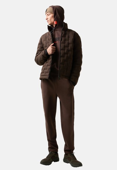 Brun pufferjakke med quiltet design, brun hoodie, matchende bukser og mørke sneakers, med en subtil logo og kontrasterende accenter.