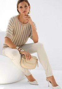 Beige gestreepte gebreide trui, roomkleurige strakke broek, witte puntige hakken. Gouden armbanden en een lichtbeige schoudertas met een klep.