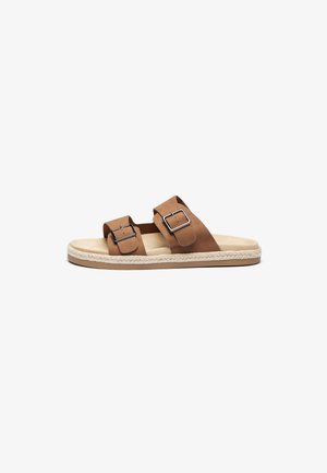 Brown suede slide sandals with two adjustable buckles, a soft beige footbed, and a woven jute sole. Flat, open design for easy wear.