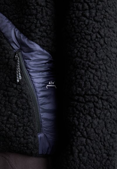 Giacca in fleece nera con fodera in contrasto blu navy, dettagli con tasche zippate e materiale esterno testurizzato. Logo parzialmente visibile vicino alla cerniera.