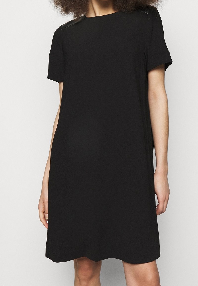 Sort kortærmet kjole med en løs pasform, der har teksturerede læderdetaljer på skuldrene og en simpel rund halsudskæring.