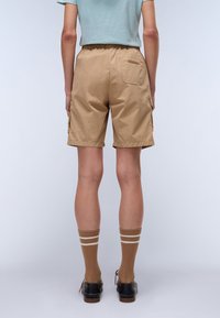 Shorts cargo beiges avec une taille élastique, des poches latérales et une poche arrière. Portés avec des chaussettes marron à rayures blanches et des chaussures noires.