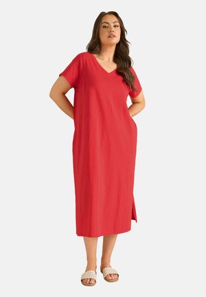 Kobieta stoi z rękami za plecami, ubrana w czerwoną midi sukienkę z krótkim rękawem i dekoltem w szpic oraz białe plecione sandały wsuwane na stopę, na białym tle.