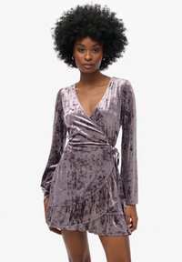 Fioletowa welurowa sukienka o kroju zawijanym z długimi rękawami, głębokim dekoltem w kształcie litery V, falbaną spódnicą i wiązaniem w talii, prezentująca delikatną, zmiażdżoną teksturę.