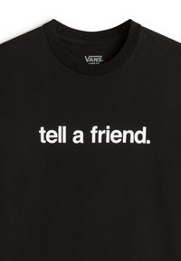 T-shirt noir Vans coupe ample avec texte blanc « tell a friend. » centré sur la poitrine.