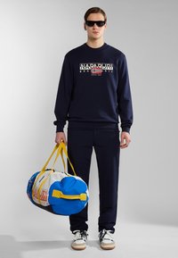Navy sweatshirt with printed graphic, matching relaxed pants, and a colorful duffel bag with blue, yellow, and white sections and handle.