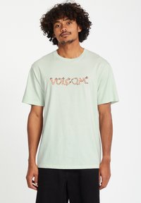 Ljusgrön bomullströja med kort ärm och rund hals, med en stiliserad "VOLCOM"-logotyp i orange och svart.