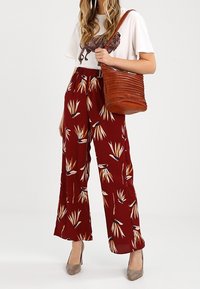 Červené široké kalhoty s květinovým vzorem, kombinované s bílým tričkem s potiskem a texturovanou hnědou kabelkou přes rameno. Šedé podpatky dotvářejí celý outfit.