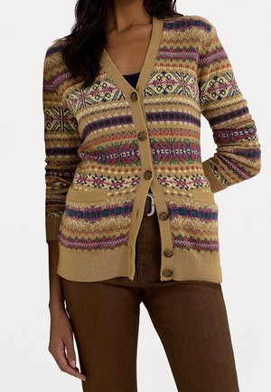 Femme portant un cardigan boutonné à motifs aux tons or, violet et vert, par-dessus un haut noir, accompagné d'un pantalon marron et d'une ceinture.