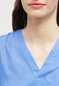 Maglietta blu da scrub con scollo a V, maniche corte e una texture liscia. Il tessuto sembra leggero e traspirante.