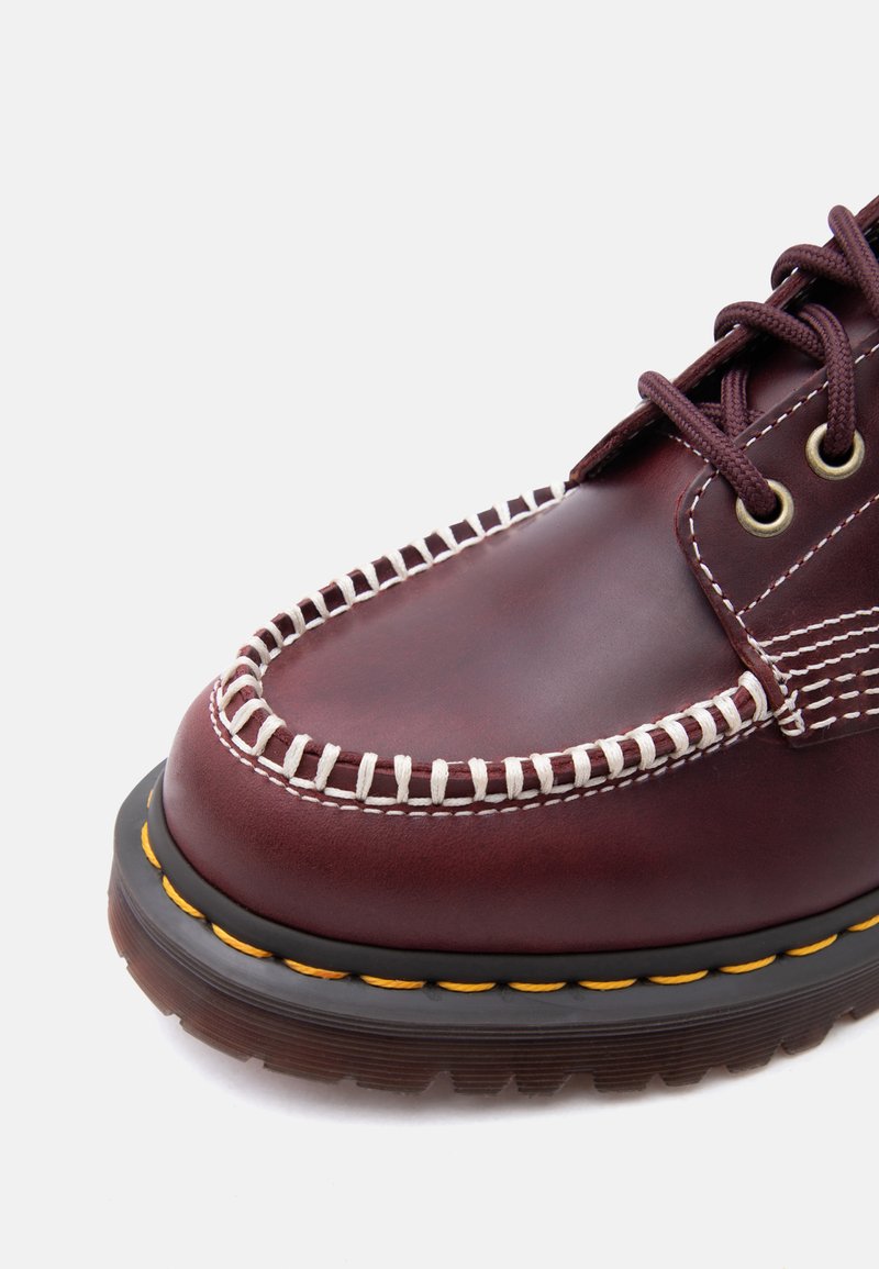 Burgundy leather moc toe shoe with white hand-stitched detailing, brown laces, metal eyelets, and thick black sole with yellow stitching.