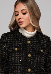 Frau mit blondem Haar, die eine schwarze strukturierte Jacke mit goldenen Knöpfen und einen weißen Rollkragenpullover trägt, blickt nach unten mit geschlossenen Augen.