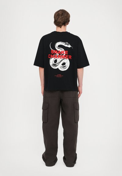 T-shirt en coton noir avec un graphique de serpent blanc et un texte rouge indiquant "SNAKE CHARMER", associé à un pantalon cargo marron foncé.
