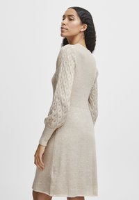 Beige gestricktes Kleid mit strukturierten, zopfgestrickten langen Ärmeln und einem taillierten Oberteil, das sich an der Taille leicht ausweitet, um eine lässige Silhouette zu schaffen.