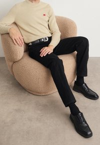 Mann, der einen beigen Pullover, schwarze Hosen mit einem Gürtel mit silberner Schnalle und schwarze Schuhe trägt, sitzt auf einem strukturierten beigen Sessel.