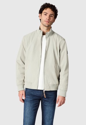 Mand iført lyse beige lynlåstrøje, hvid skjorte og blå jeans, stående mod en ensfarvet lys grå baggrund.