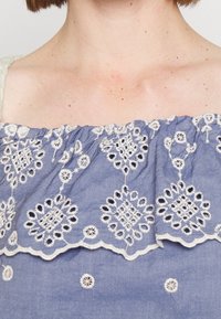 Blaues, gesticktes Oberteil mit komplizierten weißen Blumenmustern, Rüsche am Ausschnitt und weichem, leichtem Stoff mit strukturierter Oberfläche.