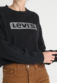 Musta collegepaita, jossa on hopeinen Levi's-logo, pyöreä kaula-aukko ja pitkät hihat, yhdistettynä ruskeisiin samettihousuihin, joissa on nappikoristelu.