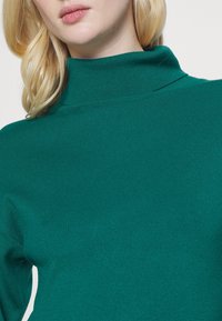Col roulé côtelé vert avec un col haut et une texture lisse, ajusté de près. Le détail au niveau de l'encolure est minimal, sans ornements supplémentaires.