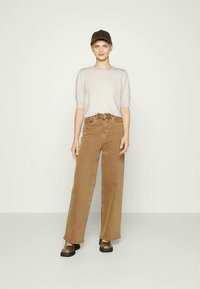 Camisola de malha curta cinza claro, combinada com calças largas de cintura alta castanhas e sapatos escuros de slip-on, acessorizada com um boné escuro.