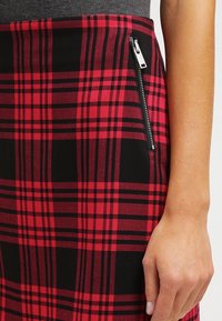 Rode en zwarte geruite rok met een gladde textuur, met een zijritssluiting en een aangesloten ontwerp. Accenten omvatten zwarte lijnen in het patroon.