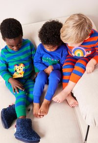 Tre børn i farverige pyjamas: grøn og blå stribet med en løve, blå med en dinosaur, og rød-orange stribet med en regnbue.