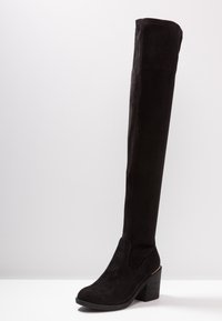 Botte noire en daim au-dessus du genou avec bout arrondi, talon bloc empilé et subtil accent doré sur le bord du talon.