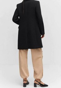 Zwarte op maat gemaakte jas met een gestructureerd ontwerp, gecombineerd met ruime beige broeken en zwarte blokhak schoenen. Gladde stof, minimalistische stijl.