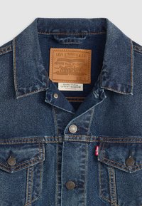 Donkerblauwe spijkerjas met een puntige kraag, bruin leren label, koperen knopen en oranje stiksels. Heeft een borstzakje. Maat M.