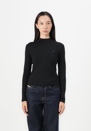 Svart langermet topp med høy hals, slim fit, med en liten logo på brystet. Bæres med mørkeblå vide jeans.