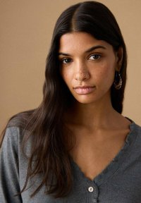 Cardigan gri cu decolteu ondulat, având o închidere cu un singur nasture. Modelul are păr lung și întunecat și poartă cercei rotunzi.