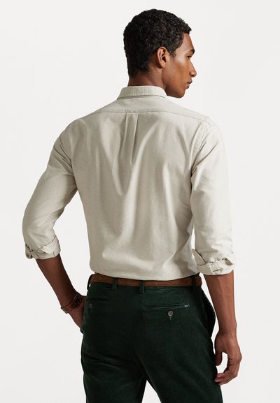 Chemise boutonnière beige clair avec des poignets retroussés, présentant un détail de passepoil au dos, associée à un pantalon en velours côtelé vert foncé.