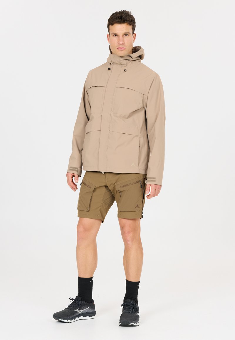 Mand stående iført en beige hættejakke, brune cargoshorts med lynlåslommer, sorte sokker og sorte atletiksko på hvid baggrund.