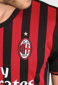 AC Milan -pelipaita, jossa on punaisia ja mustia pystysuoria raitoja, pyöreä pääntie, brodeerattu seuran logo ja kultatähti Embleemin yläpuolella.